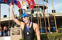 Amanda Weir im schwarzen Badeanzug und mit blauer Badekappe trägt eine Medaille um den Hals. Mit der rechten Hand winkt sie ins Publikum.