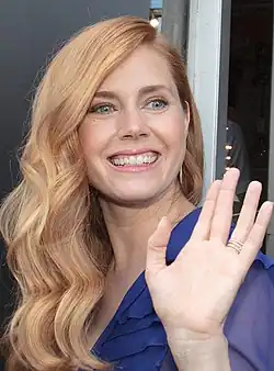 Amy Adams, eine junge Schauspielerin mit blonden langen Locken, die grinst und dem Fotografen zuwinkt.