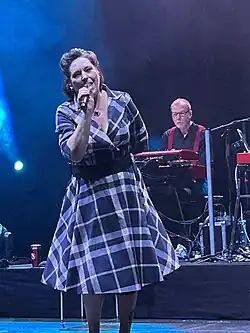 AnNa R. auf einer Bühne, die ein Mikrofon hält und in es singt, beim Abschlusskonzert ihrer König:in-Tour in Bremen 2024. Sie trägt ein kariertes Kleid mit weitem Rock und hat ein Haarband oder eine Tiara auf. Der Hintergrund ist von lila und blauen Bühnenlichtern beleuchtet, die eine rauchige Atmosphäre erzeugen. Hinter ihr ist ein Mann in schwarzem Hemd und mit roten Hosenträgern an einem roten Keyboard zu sehen. Die Momentaufnahme aus der Live-Performance wird durch die dramatische Beleuchtung und Bühnenpräsenz hervorgehoben.
