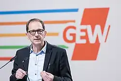 Zu sehen ist Andreas Keller, stellvertretender Vorsitzender der GEW, der an einem Mikro spricht. Im Hintergrund ist das GEW-Logo zu sehen.