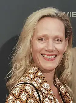 alt=Anna Schudt (2023) mit mittellangem, blondem Haar, die ein braunes Hemd mit weißen Blumenmustern und schwarzen Punkten trägt. Anna Schudt trägt auffällige, große goldene Ohrringe und eine schwarze Tasche über der Schulter. Der Hintergrund des Bildes ist dunkel, und darauf sind teils lesbare Buchstaben sichtbar, die mit „VIE“ beginnen.