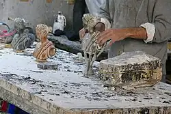 Auf einer Werkbank stehen drei Versionen eines Frauentorsos mit abgewandtem Kopf. Die hinterste, leicht unscharf, ist schwarz und eine Naht ist zu sehen. Der mittlere ist rotbraun, der vordere ist grau und ist mit Nägeln gespickt, steht auf einem trichterförmigen Sockel und ist links und rechts von Zusatzstangen (den ehemaligen Zuleitungsrohren) umgeben. Ein Mann in grauem Pulli hält diesen letzten Torso mit beiden Händen.