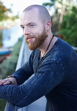 Aramis Merlin mit rasiertem Kopf und kurzem, blondem Bart und blauen Augen, trägt ein dunkelblaues Oberteil. Er lehnt mit beiden Armen auf ein Geländer, wobei mehrere Ringe an den Fingern sichtbar sind.Er lächelt. Im Hintergrund ist eine unscharfe Outdoor-Szenerie zu erkennen, die Grünflächen und möglicherweise Wasser umfasst, was darauf hindeutet, dass er sich auf einer Brücke oder einer ähnlichen Struktur befindet.