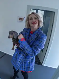 Ariane Alter (2021) lächelt und hält ihren kleinen kleinen Hund "Wolfgang". Sie trägt einen blau karierten Mantel mit einem roten Hemd darunter. Der Hund scheint ein Dackel zu sein, mit einem braun-schwarzen Fell. Im Hintergrund ist eine weiße Wand zu sehen, an der ein kleines Poster hängt. Außerdem führt eine Türöffnung in einen weiteren Raum. Das Poster an der Wand enthält ein Bild und etwas Text, der jedoch nicht klar erkennbar ist. Die Szene vermittelt eine warme und einfühlsame Stimmung, die die Verbindung zwischen der Person und dem Hund hervorhebt.