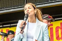 Negah Amiri, die ein Mikrofon in der Hand hält und spricht. Sie trägt einen hellblauen Blazer über einem weißen Oberteil. Im Hintergrund ist ein gelbes Banner mit roter Schrift zu sehen. Außerdem befinden sich im Hintergrund mehrere Personen, darunter eine mit einer Kappe und Sonnenbrille.