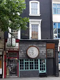 Frontale Farbfotografie von einer schmalen schwarzen Häuserfassade mit zwei oberen Stockwerken. Die Fensterumrandungen der Obergeschosse sind weiß und teilweise verziert. Im Erdgeschoss ist ein Laden mit einer riesigen Uhr zu sehen, dessen Ziffern bis 13 gehen.