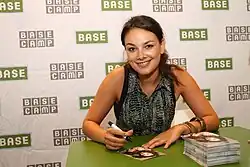 Janina Uhse bei einer Autogrammstunde BASE Camp 2012. Sie sitzt an einem Tisch, der eine grüne Oberfläche hat. Sie hält einen schwarzen Marker in der Hand und signiert ein Foto. Auf dem Tisch liegt ein Stapel Autogrammkarten mit ihrem Foto. Der Hintergrund besteht aus einem Muster mit den Worten "BASE CAMP" und "BASE", die in grünen und grauen Kästchen wiederholt erscheinen. Janina Uhse trägt ein ärmelloses, gemustertes Oberteil und mehrere Armbänder am rechten Handgelenk.