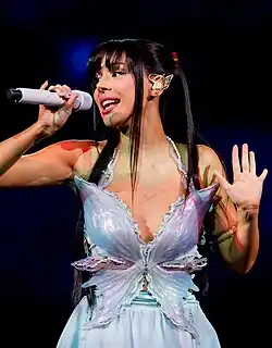 Ayliva singt auf der Bühne und hält ein Mikrofon in der linken Hand und die rechte Hand erhoben hat. Sie trägt ein schmetterlingsförmiges Kostüm mit glitzernden Details mit einem großen, schillernden Schmetterlingsdesign auf der Brust. Die Flügel des Schmetterlings sind detailliert gestaltet und scheinen aus einem glänzenden, reflektierenden Material zu bestehen. Ayliva hat langes, dunkles Haar und trägt auf der linken Seite des Kopfes eine Haarspange in Form eines Schmetterlings.