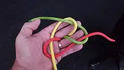 Das dritte Kardeel (gelb) führt man unter dem zweiten Ende (grün) durch und nach oben durch die Bucht des zuerst bewegten Kardeels (rot).