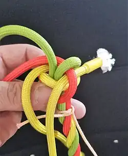 Will man die Führung oben haben, damit die Kardeele enger zur Mitte des Knotens herauskommen und die Kardeele sauberer zusammengedreht werden könnten, so legt man die zweite Führung (hier gelb) über die erste Führung (gelb), um das grüne Kardeel und durch die rote Schlaufe hindurch