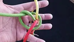 Das grüne Kardeel wird mit einem großen Törn nach hinten linksherum um den Stiel des Knoten geführt und der Tampen wird durch seine eigenen Bucht nach oben geführt. Man erhält mit dem grünen Kardeel quasi einen Überhandknoten