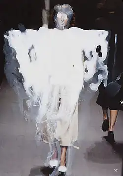 Ein Model läuft in einem Kleidungsstück über den Laufsteg, das mit weißer Farbe grob überstrichen ist, auch das Gesicht ist mit weißer Farbe verfremdet, was einen einen surrealen Effekt erzeugt.