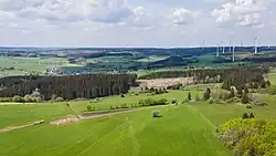 Luftbild über teils bewaldete, im Vordergrund überwiegend Wiesenflächen. Zum Hintergrund hin hügeliger, eine kleine Siedlung und am Horizont Windkraftanlagen