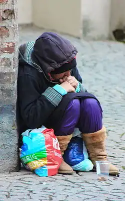 Bettlerin auf etwas Niedrigem sitzend, Einkaufstüten aus Polyethylenfolie gefüllt mit Textilien, Becher mit Münzen am Pflaster