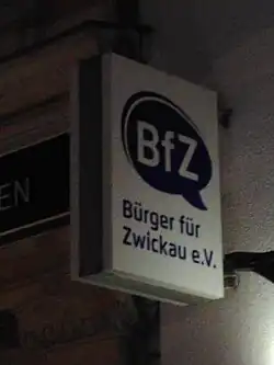 Schild an einer Hauswand. »BfZ« in weiß in einer blauen Sprechblase, daruntern »Bürger für Zwickau e.V.« in blau. Weißer Hintergrund