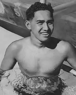 Dunkelhaariger Mann in den 20er Jahren mit nacktem Oberkörper und Badehose bekleidet in einem Schwimmbecken stehend leicht nach rechts in die Kamera lächelnd in schwarzweiß.