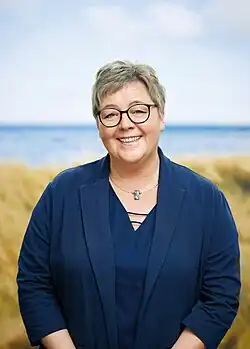 Eine Frau mit kurzen grauen Haaren und Brille, gekleidet in einem marineblauen Outfit, lächelt, während sie im Freien mit einem unscharfen Feld und Wasser im Hintergrund steht.