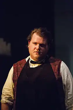 Björn Meyer trägt ein weißes Hemd mit hochgekrempelten Ärmeln, dazu eine schwarze Fliege. Über dem Hemd ist eine dunkle Schürze zu sehen, die über einem roten, gemusterten Oberteil getragen wird. Der Hintergrund ist dunkel gehalten, wodurch der Fokus auf die Person gelenkt wird. Es gibt keine zusätzlichen Objekte oder Details, die ablenken. Das Foto entstand 2015 bei einer KARLMAYSELF-Vorstellung in Hamburg.