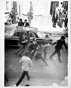 Schwarz-Weiß Foto, das im Zentrum eine schwarze Frau zeigt, die von drei Polizisten an den Armen gehalten wird und schreit. Rund um die vier sieht man weitere Menschen und ein Auto, alle Menschen im Vordergrund des Bildes scheinen in Bewegung zu sein.