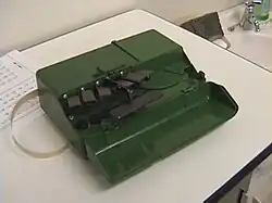 Ein kompaktes grünes Kästchen von etwa der Größe einer Zigarrenkiste. Der vordere Gehäuseteil bildet einen Deckel, er ist nach vorn heruntergeklappt und gibt den Blick auf die Bedienelemente frei: 6 schwarze Tasten für die Punkte 1 bis 6 sowie in der Mitte eine nach vorn herausstehende, breite Leertaste. Links tritt ein schmaler Papierstreifen aus dem Gehäuse heraus.