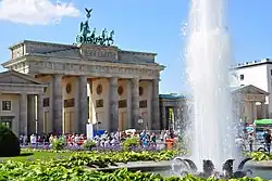 Blick auf Brandenburger Tor mit einer Grünfläche und Fontäne im Vordergrund. Das klassizistische Tor besteht aus sechs Säulen mit antiken Reliefszenen in Durchgängen, einem Giebelfeld mit antiker Szene und auf dem Dach die Quadriga in oxidiertem Grünspan.