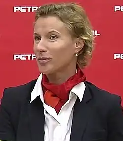 Langerholc steht vor einer roten Wand. Sie trägt eine dunkle Jacke über einem weißen Hemd sowie ein rotes Halstuch.