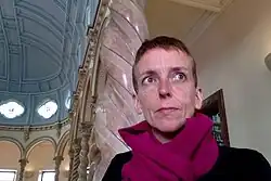 Ilaria Brocchini mit kurzen, blonden Haaren und leuchtend pinken Schal, steht in einem historischen Raum mit Säulen und hohen Decken. Sie trägt ein schwarzes Oberteil. Das Gebäude hat mehrere Säulen mit spiralförmigen Mustern bestehen aus rosa-weißen Materialien und tragen Bögen. Die gewölbte blaue Decke hat runde Fenster, die das Licht hineinlassen, und sorgt für ein himmlisches Flair. Ein Bücherregal ist im rechten Bereich des Bildes zu sehen.