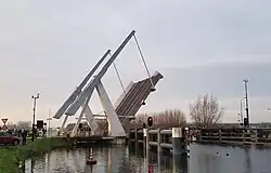 Geöffnete Brücke über ein stilles Gewässer. Die Fahrbahn ist mit einem über der Straße angebrachten, schlichten Gegengewicht aus hellgrau angestrichenem Stahl verbunden und klappt zu einer Seite auf.