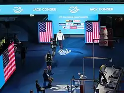 Halbtotale: Am rechten Bildrand ist das Schwimmbecken, links eine Tribüne. In der Mitte steht ein Kameramann und richtet seine Kamera auf Jack Conger, der in blauer Trainingshose, weißer Jacke und dunkler Badekappe in die Halle kommt.