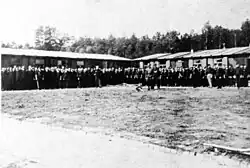 Häftlinge stehen angetreten vor den Baracken und den Wachen im SS-Arbeitslager Budzyń, vor ihnen befindet sich ein großer leerer Platz, hinter den Baracken sind Bäume zu sehen