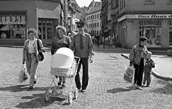 Zwei Frauen in Kleidung der 1990er Jahre und ein Mann in Uniform mit Krawatte schieben einen Kinderwagen über Kopfsteinpflaster. Im Hintergrund sind Ladengeschäfte zu sehen.