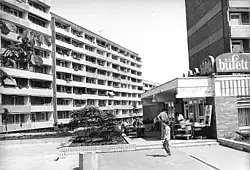 Schwarz-Weiß-Foto eines Wohnkomplexes mit mehreren Etagen und einem angrenzenden Restaurant namens "Büfett". Menschen sitzen draußen. Die städtische Szene aus dem Jahre 1990 Cottbus, Sachsendorf zeigt ein 8 stöckiges Wohnhaus. In diesem Wohnhaus im Neubaugebiet Sachsendorf-Madlow wohnte die Terroristin Susanne Albrecht alias Ingrid Jäger von 1980 bis Anfang 1985. Im Vordergrund liegt ein kleiner Platz mit einem Baum und einigen Pflanzen, die eine grüne Note hinzufügen. Links sieht man ein großes Wohngebäude mit mehreren Balkonen, auf denen Kleidung zum Trocknen aufgehängt ist, was den Eindruck eines lebendigen Wohnviertels vermittelt. Rechts im Bild ist der Eingang eines Buffet-Restaurants zu erkennen, gekennzeichnet durch ein großes Schild mit der Aufschrift „büfett“. Vor dem Restaurant stehen einige Menschen, die miteinander sprechen oder etwas kaufen. Die Kleidung der Menschen und die Architektur der Gebäude unterstreichen den zeitlichen Kontext des Bildes. Ein Mann geht über den Platz in Richtung des Restaurants, wodurch Bewegung in die Szene gebracht wird.