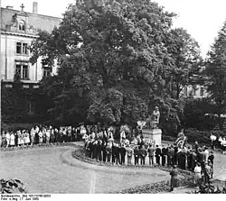 viele große Kränze am Denkmal, Männer stehen sehr ordentlich im Halbkreis darum, Frauen stehen etwas abseits. Perspektive ist schräg rechts von oben, im Hintergrund ein großer Baum und Gebäude, Schwarz-weiß-Bild