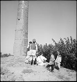 Links im Bild ragt ein Minarett vor einem freien Himmel nach oben aus dem Bild heraus. Rechts davon ist Buschwerk. Davor ist eine Gruppe von Menschen in afghanischer Kleidung zu sehen.