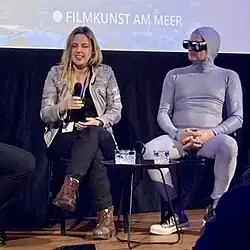 Camilla Guttner sitzt mit einer weiteren Person auf einer Bühne. Sie hält ein Mikrofon und scheint zu sprechen. Sie trägt eine graue Jacke, eine schwarze Hose und braune Stiefel. Die Person auf der rechten Seite ist vollständig in einen eng anliegenden grauen Ganzkörperanzug gekleidet, einschließlich einer Kapuze, die den gesamten Kopf bedeckt. Sie trägt weiße Turnschuhe.Zwischen den beiden Personen befindet sich ein kleiner Tisch mit zwei Gläsern Wasser darauf. Im Hintergrund ist eine Leinwand sichtbar, auf der der Text „FILMKUNST AM MEER“ steht.