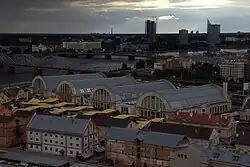 Farbfotografie in der Obersicht von vier großen Hallen im Rundbau mit vielen umliegenden Gebäuden. Eine lange Grünfläche erstreckt sich im vorderen Bereich. Im linken Hintergrund ist ein brauner Wolkenkratzer und in der hinteren Mitte geht eine Flussabzweigung vom rechten großen Fluss. Rechts verbindet eine Brücke das hintere Ufer, auf dem ein riesiger, moderner Turm steht, mit dem vorderen Ufer.