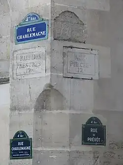 Heutiges Straßenschild (doppelt), dazwischen der frühere Name der Straße „Rue des Prestres“