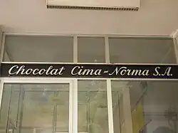 Dieses Bild zeigt einen kursiv gestalteten Schriftzug der Chocolat Cima-Norma S.A. (weiss auf dunklem Grund), der über einer Glastür angebracht ist.