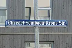 Blaues Straßenschild mit weißer Schrift vor einer braungrauen Holzfassade