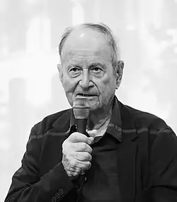 Schwarzweiß-Porträt eines älteren Mannes mit Mikrofon in der Hand vor hellem Hintergrund