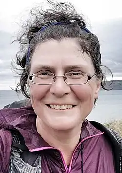 Christine Thürmer trägt eine lilafarbene Jacke und schaut lächelnd in die Kamera. Ihr dunkles lockiges Haar ist zurückgebunden. Sie trägt eine Brille und im Hintergrund ist ein See zu sehen.