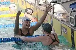 Cierra Runge ist von vorn zu sehen, sie trägt eine weiße Bademütze und hat die Schwimmbrille auf die Stirn hochgeschoben. Beide Arme sind erhoben, um die ihr entgegenkommende Missy Franklin zu umarmen.
