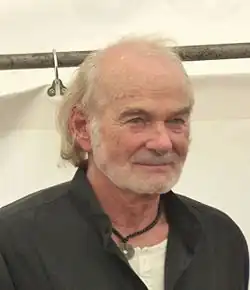 Claus Dieter Clausnitzer mit hellem Haar und Halbglatze. Er trägt ein dunkles Oberteil über einem weißen T-Shirt und eine Halskette mit einem runden Anhänger. Im Hintergrund sind eine Metallstange mit einem Haken daran sowie ein weißes Tuch oder Vorhang sichtbar, das von der Stange hängt. Die Umgebung ist schlicht gehalten, ohne auffällige Dekorationen oder Farbakzente.