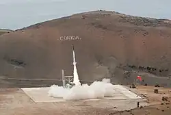 Startsequenz mit Rauchentwicklung am Boden der abhebenden Rakete vor einem Bergpanorama.