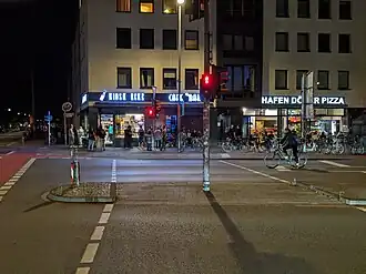Junge Menschen cornern an einer Straßenecke im Hansaviertel in Münster. An der Straße befinden sich auch ein Kiosk und eine Imbissstube.