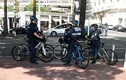 Fünf Polizisten auf Mountainbikes, alle mit Sonnenbrille, aber nur vier der fünf mit Fahrradhelm, stehen in städtischer Umgebung auf dem Gehweg vor einem Zebrastreifen. Alle sind mit dunkelblauen Uniformen der Police municipale bekleidet: schwarze Stiefel, lange Hosen, Windjacke und Kopfbedeckung. Im Hintergrund befindet sich ein Juweliergeschäft der Kette „Cartier“.