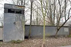 links ein grauer Wachturm mit abblätterndem Putz an einer circa 2 Meter hohen grauen Mauer, rechts ein aufgemaltes weißes Schild
