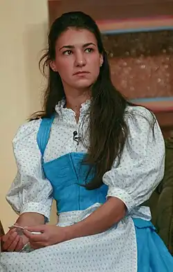 Rosetta Pedone trägt traditionelle Kleidung, die an historische Designs erinnert. Sie hat eine weiße Bluse mit kleinen blauen Mustern und puffigen Ärmeln an. Die Bluse hat einen hochstehenden Kragen mit gerüschten Details. Darüber trägt sie ein blaues Schürzenkleid, das vorne mit einem Gürtel oder Bund gerafft ist und einen taillierten Schnitt aufweist. In den Händen hält die Person einen Zettel und blickt nachdenklich in die Ferne. Der Hintergrund zeigt eine strukturiert wirkende Wand mit einer Art Regal oder Vorsprung darauf. Der Gesamteindruck des Bildes erinnert an eine sorgfältig gestaltete Kostümierung, die für ein Theaterstück genutzt werden könnte.