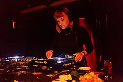 Ostbam steht an DJ-Player, das Licht ist sehr gedämpft und orange und dunkel. Sie hat halblanges Haar, trägt Kopfhörer, einen dunklen Pullover und eine Kette.