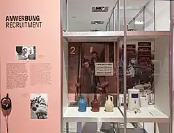 Zu sehen ist ein Ausstellungselement der Ausstellung „Wer wir sind. Fragen an ein Einwanderungsland“ im Kapitel „Anwerbung“. Links ist ein beschreibender Text. In einer gläsernen Vitrine stehen drei Trinkgefäße, die sog. Gastarbeiter erhalten haben auf ihrer Einreise nach Deutschland. Rechts ist ein Lungenvolumenmessgerät zu sehen, das für Gesundheitsuntersuchungen im Rahmen der Anwerbeabkommen verwendet wurde. Im Hintergrund sind historische Fotoaufnahmen zu erkennen. Die Objekte sind Teil der DOMiD Sammlung. DOMiD wirkte als Kooperationspartner an der Ausstellung in der Bundeskunsthalle Bonn mit.
